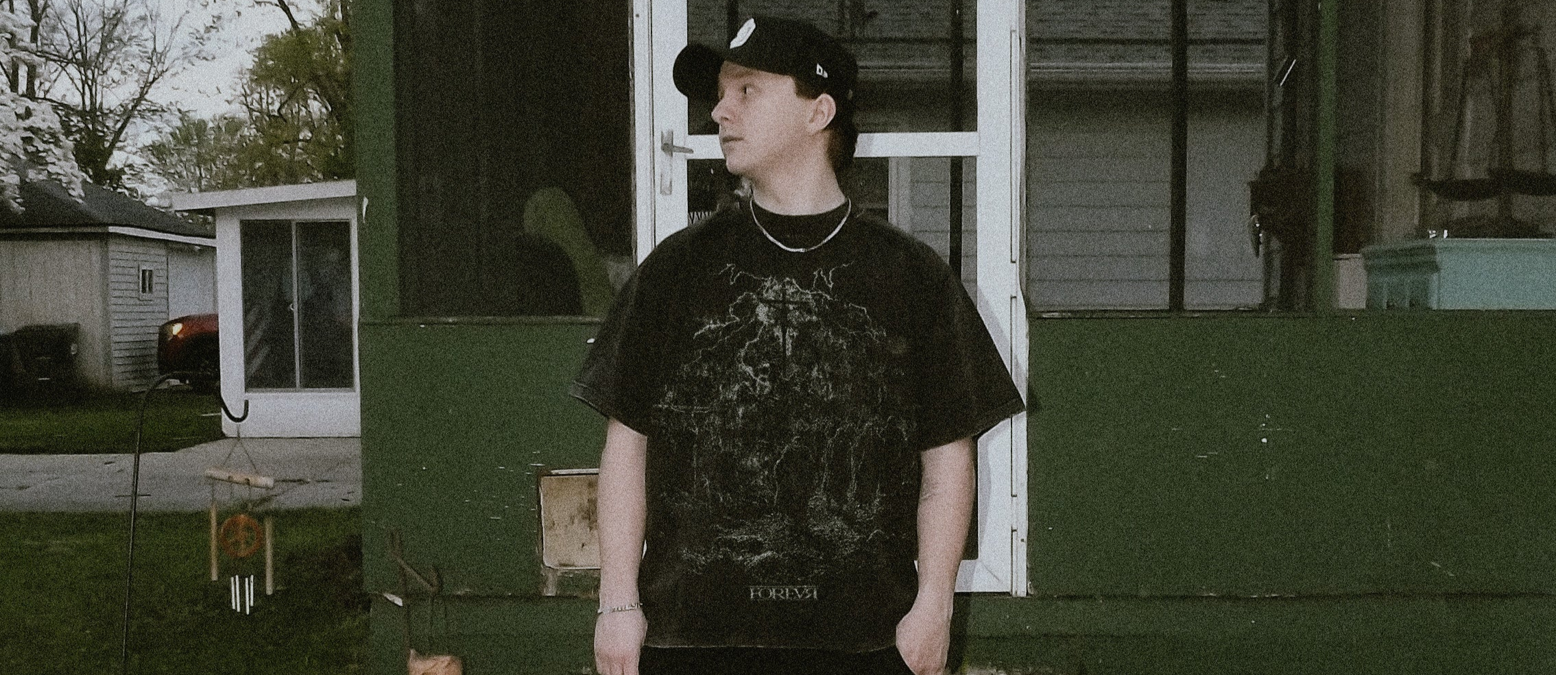 Person wearing a black t-shirt with text and dark pants standing in front of a house.